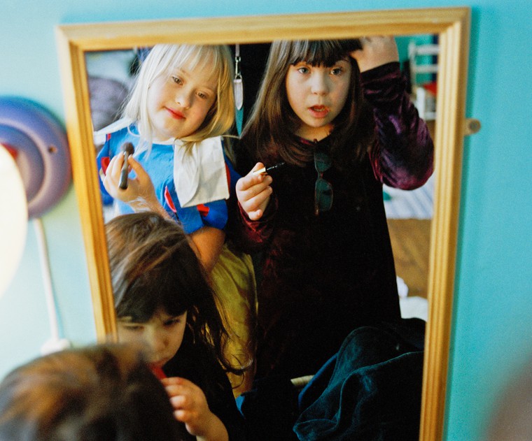 A young girl with Down syndrome smiles at herself in the mirror, wearing a dress-up costume and holding a makeup brush. The child is playing with two young children without visible disabilities. 